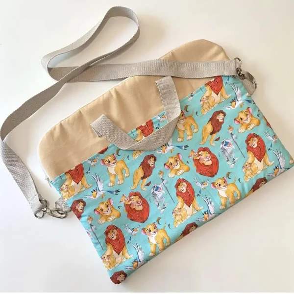 Funda para portátil hecha a mano en tela beige y estampado infantil de personajes sobre fondo turquesa, con asas cortas y bandolera desmontable en gris claro.
