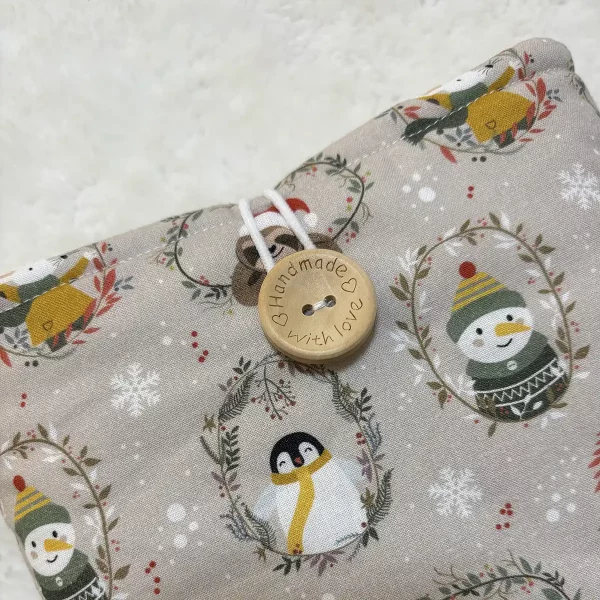 Detalle de funda acolchada para Kindle, hecha a mano en algodón beige con estampado invernal, cierre con botón de madera grabado con la frase “Handmade with love” y cordón blanco elástico.