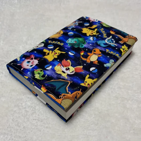 Funda artesanal de tela ajustada a un libro, vista lateral sobre superficie blanca, con elástico blanco visible en el borde.