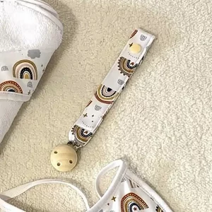 Portachupete de tela con estampado de arcoíris en tonos tierra, con clip de madera natural, sobre fondo de manta beige