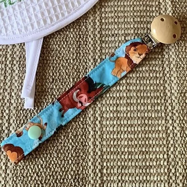 Portachupete de tela con estampado infantil de personajes sobre fondo azul celeste, clip de madera natural, sobre alfombra de yute.