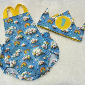 Conjunto de ranita y corona infantil en tela azul con estampado de nubes, arcoíris y confeti, tirantes y detalles en amarillo, sobre fondo blanco esponjoso.