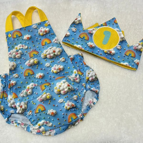 Conjunto de ranita y corona infantil en tela azul con estampado de nubes, arcoíris y confeti, tirantes y detalles en amarillo, sobre fondo blanco esponjoso.