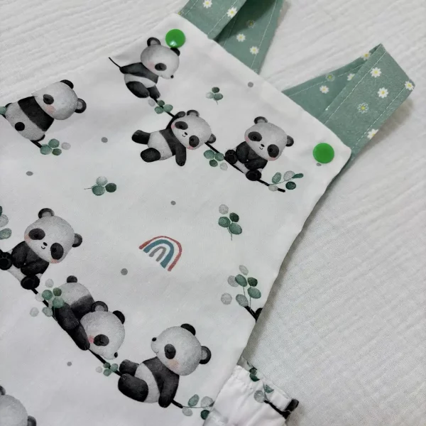Detalle de ranita de bebé artesanal con tirantes cruzados verdes y botones a presión verdes, confeccionada en algodón blanco.