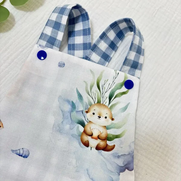 Detalle superior de ranita de bebé hecha a mano con tirantes de cuadros azules y botón a presión azul, confeccionada en algodón suave.