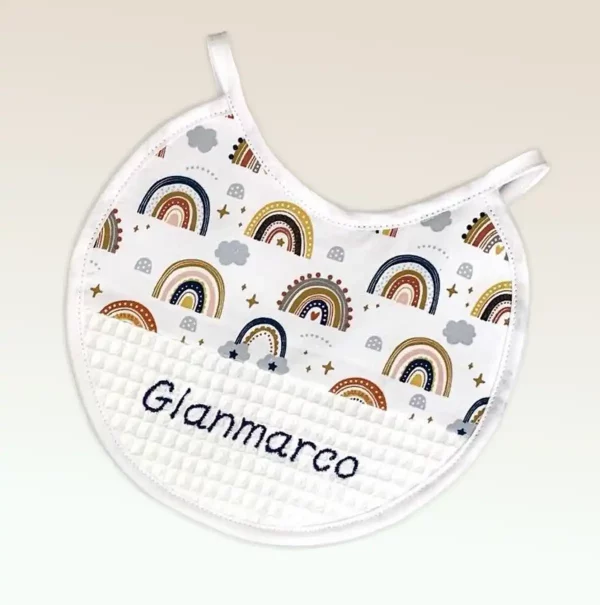 Babero personalizado de algodón con estampado de arcoíris y nombre bordado en hilo azul, ideal para bebés.